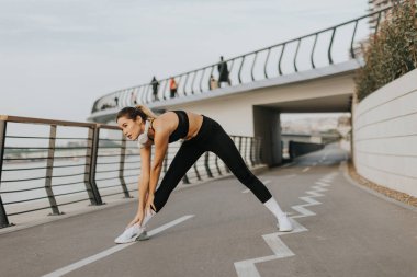 Atletik kıyafetler giyen fit bir kadın nehir kenarındaki patikada sakin su manzaralı ve sakin bir sabahın tadını çıkaran yürüyüşçülerle esneme hareketleri yapıyor..