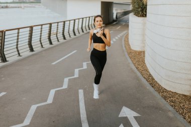 Sportif kıyafetler giymiş bir kadın, su kenarında bir patika boyunca mutlu bir şekilde koşar. Kulaklıklarıyla, canlı sabah atmosferini ve serin esintiyi kucaklıyor..