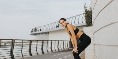 Yoğun bir koşudan sonra, genç bir kadın nefes almak için durur, canlı bir şehir ortamında kulaklıklarıyla müzik dinlerken gülümseyerek..