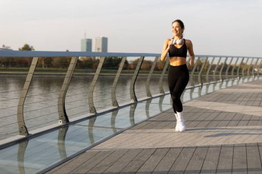 Güneş sakin suyun üzerinde yükselirken, kararlı genç bir kadın güzel bir sahil yolunda koşar, doğanın içinde zindelik ve refahını gösterir..