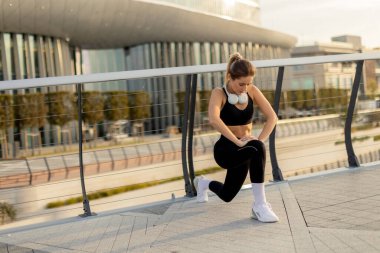 Altın saat boyunca sakin bir şehir ortamında, genç bir kadın bacaklarını düşünceli bir şekilde açar, güneş arkasında batarken zindelik ve sağlığa olan bağlılığını somutlaştırır..