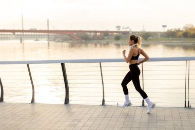 Kararlı bir koşucu, sakin sular ve şafağın yumuşak renkleri ile çevrili manzaralı nehir kenarındaki yürüyüş yolunda yürümeye devam eder. Sabahın erken saatlerindeki sakinliği kucaklar..