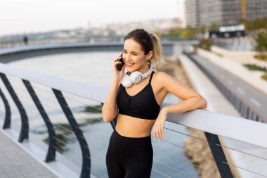 Bir koşucu nehir kenarındaki telefonunda antreman kıyafetleri ve kulaklıklar takarak sohbet etmek için durur. Sıcak güneş ışığını ve etrafındaki sakin atmosferi kucaklar..