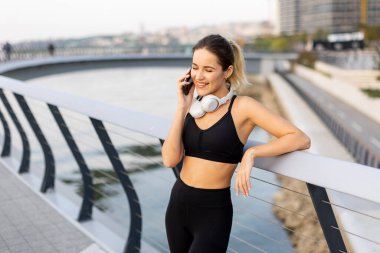 Bir koşucu nehir kenarındaki telefonunda antreman kıyafetleri ve kulaklıklar takarak sohbet etmek için durur. Sıcak güneş ışığını ve etrafındaki sakin atmosferi kucaklar..