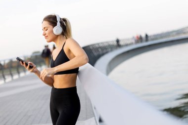 Genç bir kadın, bir köprüde, kulaklıkları takmış, günbatımının hafif parıltısında suyun kenarında günbatımının tadını çıkarırken müziğin tadını çıkarıyor..