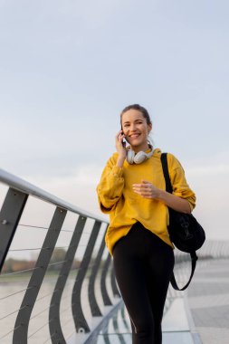 Genç bir kadın gösterişli bir yaya köprüsünde yürürken telefonuyla konuşuyor. Sakin sular, alacakaranlık yaklaşırken kentsel bir ufku yansıtıyor.