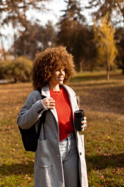 Kıvırcık saçlı neşeli bir kadın güneşli bir parkta geziyor. Renkli sonbahar yapraklarıyla süslenmiş, şık bir kıyafet giyerken elinde sıcak bir fincan kahve tutuyor..