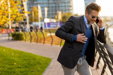 Şık günlük kıyafetler giymiş bir adam, şehir manzarasının yanında duruyor, sonbahar yaprakları ılık güneş ışığında etrafında dönerken bir telefon görüşmesi yapıyor..