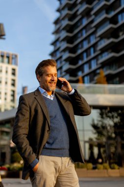 İyi giyimli bir adam, çağdaş mimariyi ve yeşilliği parlak güneş ışığı altında geçerek canlı bir telefon görüşmesi yaparak güvenli bir şekilde şehir ortamında dolaşır..