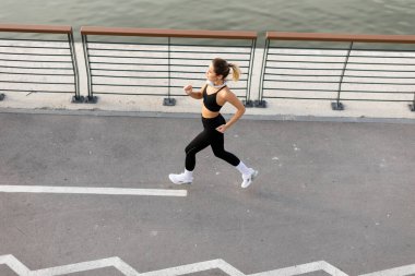 Aktif kıyafetli bir kişi sakin suyun yanında iyi korunmuş bir yolda koşuyor, şehirde enerjik bir sabah rutininin özünü yakalıyor..