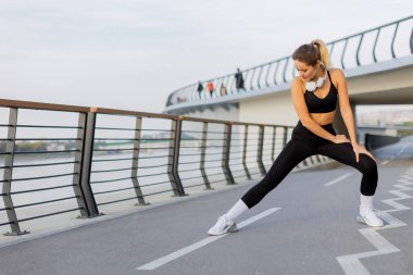 Atletik kıyafetler giyen fit bir kadın nehir kenarındaki patikada sakin su manzaralı ve sakin bir sabahın tadını çıkaran yürüyüşçülerle esneme hareketleri yapıyor..