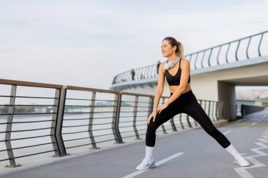 Atletik kıyafetler giyen fit bir kadın nehir kenarındaki patikada sakin su manzaralı ve sakin bir sabahın tadını çıkaran yürüyüşçülerle esneme hareketleri yapıyor..