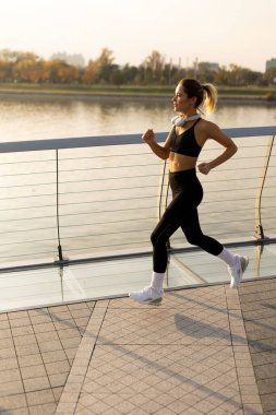 With the sun rising over the calm water, a determined young woman jogs along a beautiful boardwalk, showcasing her commitment to fitness and well-being amidst nature. clipart