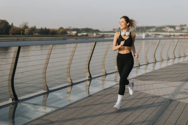 Atletik giyinmiş genç bir kadın nehir kenarında enerjik bir şekilde koşuyor, güzel bir sabahın dingin atmosferini kucaklıyor, en sevdiği müzikle besleniyor..