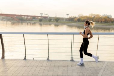 Kararlı bir koşucu nehir kenarında güvenli adımlar atar, sakin bir sabah havasını kucaklar, şehir silueti ve sonbahar ağaçları resim gibi bir zemin oluşturur..