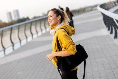 Nehir kenarındaki yolda, neşeyle sarı kapüşonlu bir kadın yürüyor. Akşam gökyüzü ılık bir parıltı yayar, sakin bir atmosfer yaratır, sakin bir gezinti için.