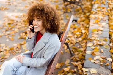 Kıvırcık saçlı genç bir kadın telefonda konuşurken neşeli bir şekilde gülüyor. Parktaki bir bankta oturuyor, canlı sonbahar yapraklarıyla kaplı.