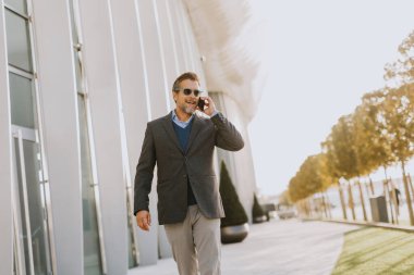 İyi giyimli bir adam, çağdaş mimari ve yeşilliklerle çevrili, öğleden sonra güneşinin sıcacık ışıltısı altında telefonuyla sohbet ederek, gösterişli bir patikada yürüyor..