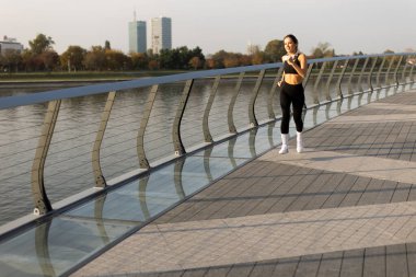 Sabah dinginliğini kucaklayan tek bir koşucu, ağaçlarla çevrili güzel bir rıhtım yolu boyunca koşuyor, şehrin ufku sakin sulara yansıyor..