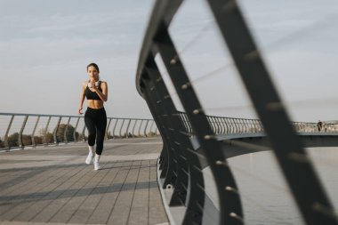 Atletik giyinmiş genç bir kadın nehir kenarında enerjik bir şekilde koşuyor, güzel bir sabahın dingin atmosferini kucaklıyor, en sevdiği müzikle besleniyor..
