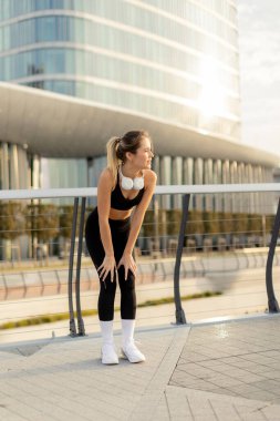 Enerjik bir egzersizden sonra, şık spor kıyafetleri içindeki genç bir kadın nefes almak için duruyor, öğleden sonra güneşinin sıcacık parıltısında nefes kesici bir şehir zeminine karşı güneşleniyor..