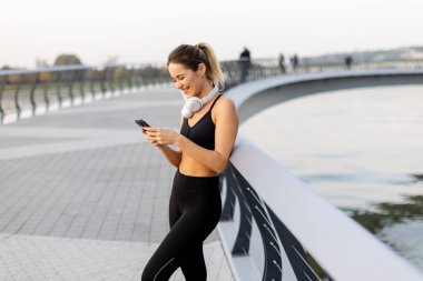 Bir koşucu nehir kenarındaki telefonunda antreman kıyafetleri ve kulaklıklar takarak sohbet etmek için durur. Sıcak güneş ışığını ve etrafındaki sakin atmosferi kucaklar..