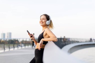 Genç bir kadın, bir köprüde, kulaklıkları takmış, günbatımının hafif parıltısında suyun kenarında günbatımının tadını çıkarırken müziğin tadını çıkarıyor..