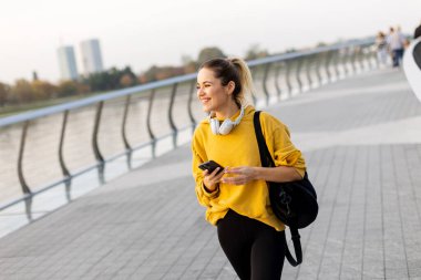 Gülümseyen genç bir kadın sonbaharda nehir kenarında yürür, rahat sarı bir kazak ve kulaklık giyer. Sakin atmosfer onun neşeli anını yakalıyor..