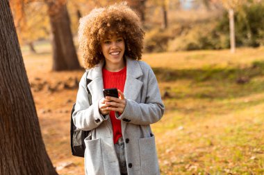 Aydınlık bir sonbahar gününde neşeli bir kadın, canlı yapraklarla çevriliyken telefonuyla bir ağacın yanında durur. Samimi kıyafeti mevsimsel renklerle çok uyumsuz..