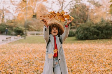 Kıvırcık saçlı neşeli bir birey sonbaharda bir parkta oynar, neşe içinde düşen yapraklardan oluşan altın bir battaniyenin arasına canlı yapraklar atar..