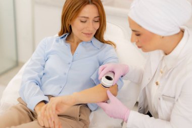 A woman sits comfortably in a modern clinic while a practitioner applies a dermatoscope device to her arm, showcasing a blend of care and advanced technology for beauty enhancement. clipart