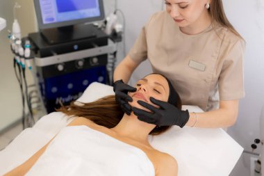 Huzurlu bir spa ortamında huzurlu bir yüz tedavisinden hoşlanan bir kadın, sakinleştirici renkler ve canlandırıcı bir deneyim sunan nazik ellerle çevrilidir..