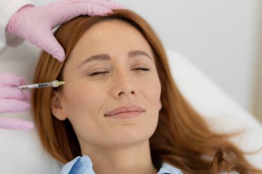 Müşteri, parlak bir klinikte rahat bir şekilde oturur ve bir profesyonel olarak cilt dolgusu tedavisi uygularken gülümser. Sakin atmosfer rahatlamayı ve gençleşmeyi destekler..