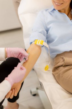 Bir sağlık uzmanı kan almaya hazırlanırken bir hasta rahat bir şekilde oturur. Ortam, rahatlama ve güveni destekleyen temiz ve parlak bir tıbbi klinik..