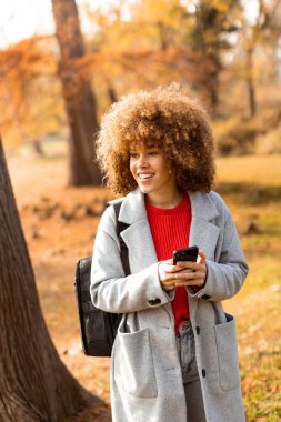 Aydınlık bir sonbahar gününde neşeli bir kadın, canlı yapraklarla çevriliyken telefonuyla bir ağacın yanında durur. Samimi kıyafeti mevsimsel renklerle çok uyumsuz..
