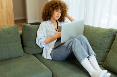 Lüks bir kanepede rahat bir şekilde oturan genç bir kadın, dizüstü bilgisayarıyla çalışırken sıcak kahvesinin tadını çıkarıyor, şık bir yaşam alanının yumuşak doğal ışığıyla aydınlatılıyor.