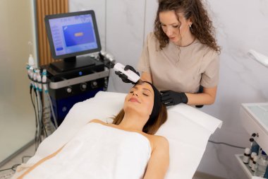 Becerikli bir terapist müşterinin alnına ultrasonik yüz masajı yapar. Rahatlama ve cilt bakımını iyileştirir..