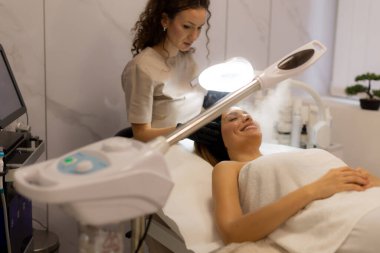 Huzurlu bir spa ortamında yüzü sisle kaplanmış bir kadın yüz tedavisiyle eğleniyor. Atmosfer rahatlama ve cilt tazelemeyi teşvik ediyor..