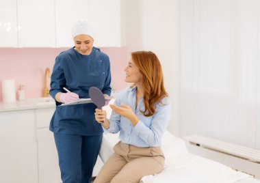 Parlak bir güzellik kliniğinde, yetenekli bir uzman, bir müşteriyle muayene masasında rahatça oturup ayna tutarken cilt bakımı seçeneklerini tartışır..