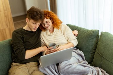 Yumuşak bir battaniyeye sarılmış bir çift yeşil bir kanepeye oturur, kahkahaları ve sıcaklığı paylaşırlar, dizüstü bilgisayarlarında gezinirler, rahat bir kupadan yudumlarlar, keyifli anılar yaşarlar..