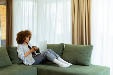 Lüks bir kanepede rahat bir şekilde oturan genç bir kadın, dizüstü bilgisayarıyla çalışırken sıcak kahvesinin tadını çıkarıyor, şık bir yaşam alanının yumuşak doğal ışığıyla aydınlatılıyor.