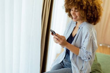 Parlak bir gülümsemeyle, genç bir kadın yeşil bir kanepede oturuyor, akıllı telefonuyla meşgul, doğal ışıkla dolu sıcak ve davetkar bir evin içinde..