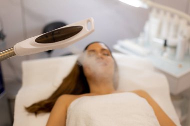 Huzurlu bir spa ortamında yüzü sisle kaplanmış bir kadın yüz tedavisiyle eğleniyor. Atmosfer rahatlama ve cilt tazelemeyi teşvik ediyor..