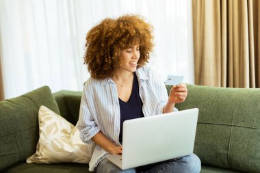 Kıvırcık saçlı genç bir kadın elinde kredi kartıyla internetten alışveriş yapıyor. Sıcak güneş ışığıyla dolu şık bir oturma odasında rahat bir koltukta oturuyor..