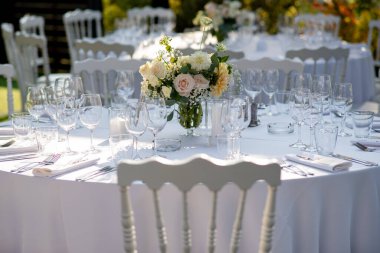 Zarif güllerin ve yeşilliklerin bulunduğu çarpıcı bir çiçek aranjmanı el değmemiş bir masanın üzerinde duruyor ve alacakaranlıktaki düğün töreninin romantik atmosferini güzelleştiriyor..