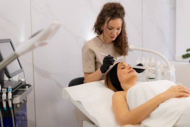 Huzurlu bir spa odasında, yetenekli bir estetisyen, rahat bir müşteriye retinol serumu uygular, konforlu bir seans sırasında cildini gençleştirmeye odaklanır..