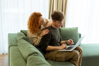 İki arkadaş evlerinde neşeli bir an yaşıyor, yeşil bir koltukta rahatça oturuyorlar, kahkaha ve neşe dolu bir dizüstü bilgisayarla işbirliği yapıyorlar..