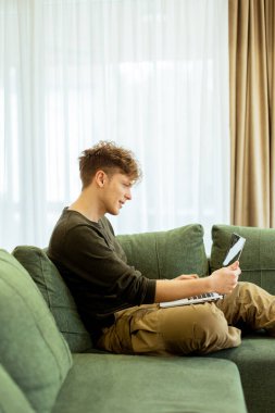Genç bir adam yeşil bir kanepede rahatça oturur, dizüstü bilgisayarına derinden odaklanır. Güneş ışığı geniş pencerelerden süzülerek davet edilen boşlukta sıcak bir atmosfer yaratır..