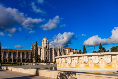 Ziyaretçiler Portekiz, Lizbon 'daki Jeronimos Manastırı' nın güzel topraklarında geziyorlar, büyüleyici mimarisi ve tüylü bulutlarla süslenmiş parlak mavi gökyüzü ile eğleniyorlar..