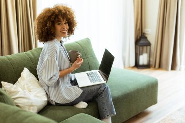 Yeşil bir kanepede rahat bir şekilde oturan genç bir kadın, doğal ışıkla dolu, parlak ve sakin bir oturma odasında laptopuyla çalışırken sıcak bir fincan kahvenin tadını çıkarıyor..